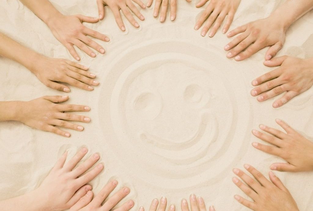 Viele Hände liegen im Kreis im Sand in der Mitte ein lachender Smiley